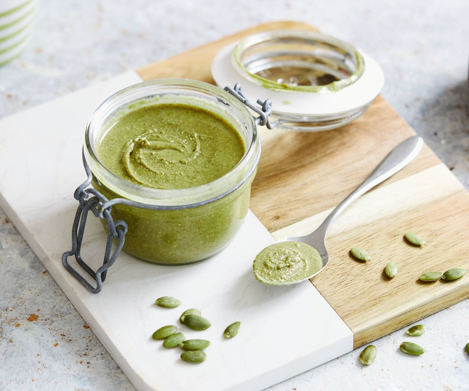 A jar of vibrant green pesto made from pepitas (hulled pumpkin seeds) sits on a counter next to a pile of the raw seeds.