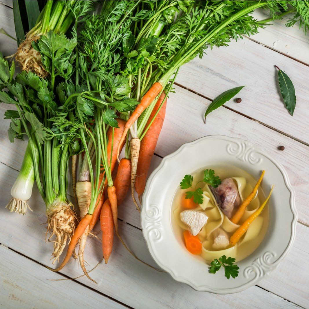 Freshly harvested carrots, celery root, and leeks arranged next to a bowl of wholesome vegetable noodle soup, showcasing garden-to-table ingredients